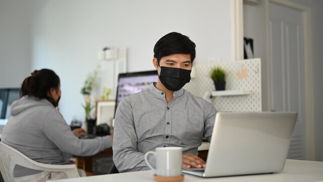 A young man freelance photographer with face mask edit photos on computer laptop at the office.