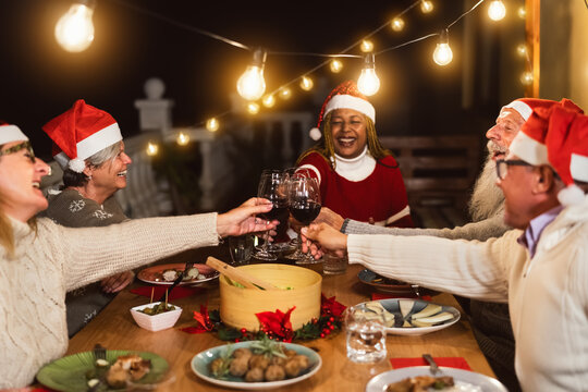 Happy multiracial senior friends toasting with red wine glasses during Christmas holidays dinner celebration on patio house party