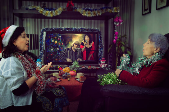 A caregiver and an old woman sitting in front of a TV screen on Christmas Eve. They sing along to a couple singers on the holiday channel. The room is decorated with tinsels.