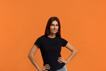 Obraz premium Yes, i will win. Are you with me. Smiling caucasian woman in black t-shirt standing with arms on belt, bit a lip and looking at camera, showing their confident. Isolated over an orange background.