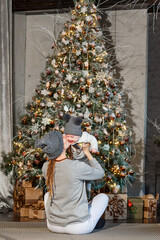 A mother kisses a smiling baby near a Christmas tree. photo shoot mother and toddler. people in gray clothes.