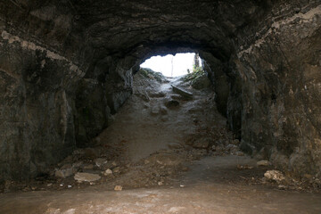 exit from cave, with natural stone walls. Exit from mountain to sunlight that illuminates trees and nature. Natural tunnel overlooking landscape. Trees and plants, framed from exit from stone adit.