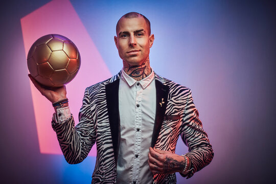 Positive and succesful football trainer dressed in custom suit with jewellery poses with golden ball in abstract light background.