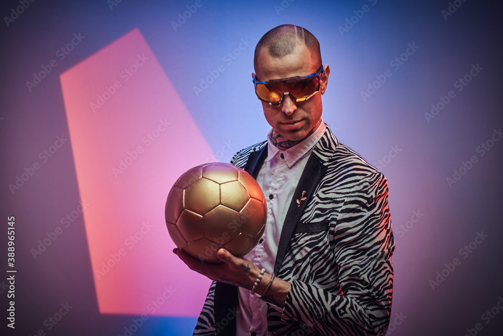 Wall mural Styled and futuristic guy with eyewears poses with golden soccer ball in abstract background.