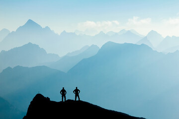 Two Men reaching summit after climbing and hiking enjoying freedom and looking towards mountains silhouettes panorama during sunrise. - obrazy, fototapety, plakaty