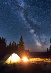 Night summer camping in forest. Back view male tourist raising hands near bright campfire and illuminated tent, enjoying beautiful dark starry sky and Milky way. Concept of tourism, camping.