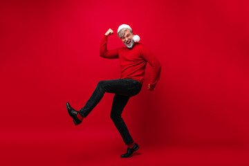 Fototapeta na wymiar Full-lenght portrait of white-haired grandpa enjoying christmas time. Funny old male model dancing on red background at new year party.