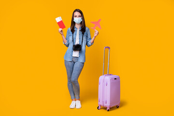 Full body photo of pretty young lady rolling bag photographer journalist travel airport quarantine show registration tickets wear facial mask casual clothes isolated yellow color background