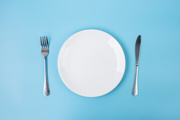 empty white ceramics plate with knife and fork on blue background. dining and kitchenware concept