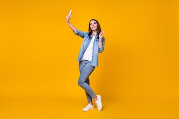 Fototapeta na wymiar Full length photo of attractive funny lady good mood hold telephone making selfies youth blog show v-sign symbol wear casual denim shirt sneakers isolated vivid yellow color background