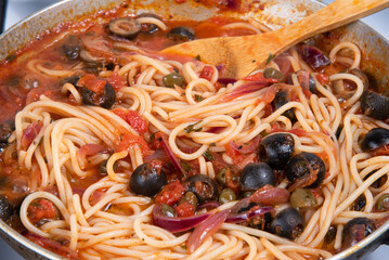 Cooking spaghetti alla vesuviana, pasta with sauce and tomatoes, olives, onions and capers - cooked al dente spaghetti with onion sauce, chopped tomatoes, olives and capers in a pan close-up