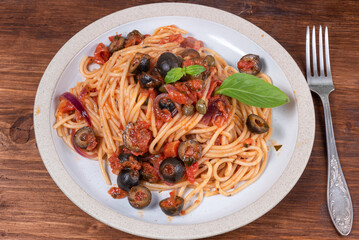 Spaghetti alla vesuviana, pasta with sauce and tomatoes, olives, onions and capers in a plate on a rustic wooden table - italian lunch