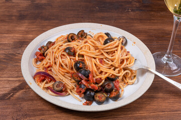 Spaghetti alla vesuviana, pasta with sauce and tomatoes, olives, onions and capers in a plate on a rustic wooden table and a glass of wine - italian lunch