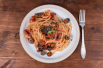 Spaghetti alla vesuviana, vegetarian pasta with sauce and tomatoes, olives, onions and capers in a plate on a rustic wooden table, top view
