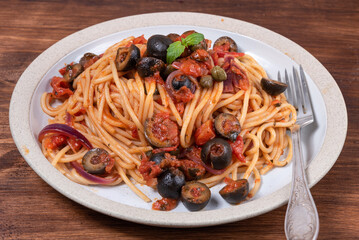 Spaghetti alla vesuviana, vegetarian pasta with sauce and tomatoes, olives, onions and capers in a plate on a rustic wooden table, top view, close-up