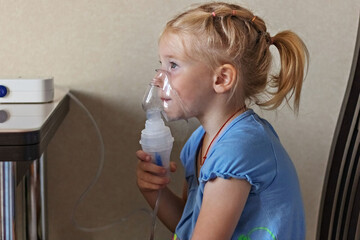 Little girl makes inhalation with a nebulizer at home, sitting on a chair. Portable apparatus for...