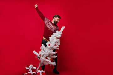Happy bearded man in knitted sweater fooling around in new year. Studio portrait of joyful white guy dancing on red background with christmas tree.