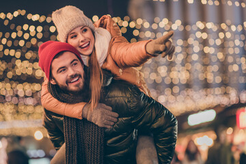 Photo of two friends students guy hug piggyback girl point finger under evening x-mas illumination outdoors