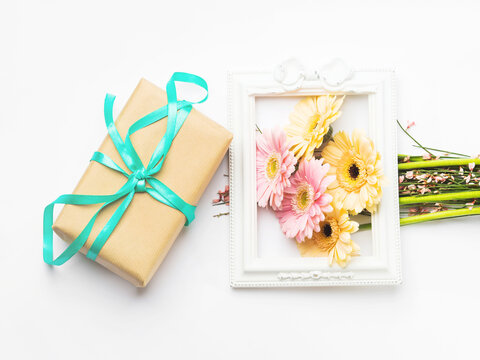Beautiful pastel pink and orange gerbera bouquet in picture frame with wrapped gift box on white background. Giving flowers for womens, mothers day.