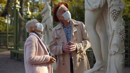 Senior couple in safety mask walking in city park looking at sculpture