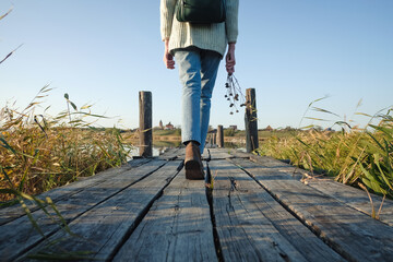 Unrecognizable woman traveler walking on old wooden bridge with bouquet dried flowers.