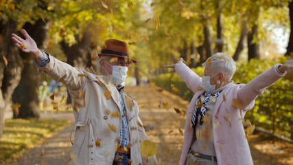 Fototapeta premium Happy aged couple in safety mask throwing fall leaves, walking and having fun in park