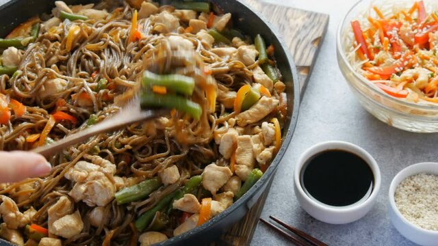 Japanese soba rice noodles with pieces of chicken, carrot, green shoots, sesame seeds, soy sauce. National food in a restaurant in large frying pan next to wooden or bamboo fork. Glass noodle salad.