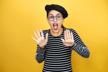 Fototapeta na wymiar Young beautiful brunette woman wearing french beret and glasses over yellow background afraid and terrified with fear expression stop gesture with hands, shouting in shock. Panic concept.