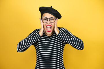 Young beautiful brunette woman wearing french beret and glasses over yellow background crazy and scared with hands on head, afraid and surprised of shock