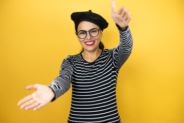 Young beautiful brunette woman wearing french beret and glasses over yellow background looking at the camera smiling with open arms for hug