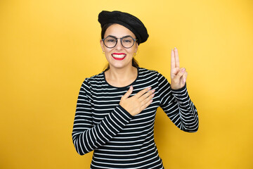 Young beautiful brunette woman wearing french beret and glasses over yellow background smiling...