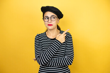 Young beautiful brunette woman wearing french beret and glasses over yellow background smiling happy pointing with hand and finger to the side