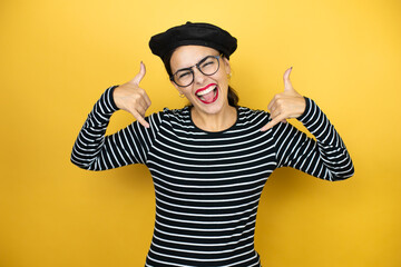 Young beautiful brunette woman wearing french beret and glasses over yellow background shouting with crazy expression doing rock symbol with hands up