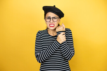 Young beautiful brunette woman wearing french beret and glasses over yellow background suffering pain on hands and fingers, arthritis inflammation
