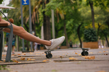 Girl sitting on a bench in the middle of the pedestrian street and putting her feet on the skateboard with a blur background of green trees.