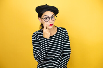 Young beautiful brunette woman wearing french beret and glasses over yellow background Pointing to the eye watching you gesture, suspicious expression