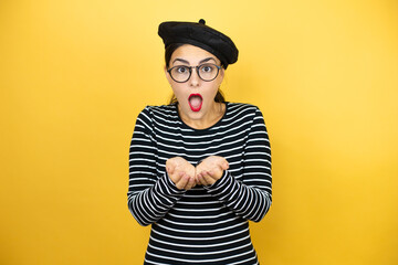 Young beautiful brunette woman wearing french beret and glasses over yellow background Smiling with hands palms together receiving or giving gesture. Hold and protection
