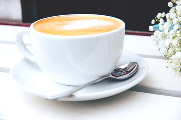 Cappuccino in a white ceramic cup and bouquet of small white flowers on a wooden bench. Open-air rustic morning coffee. Gypsophila flowers. Side view