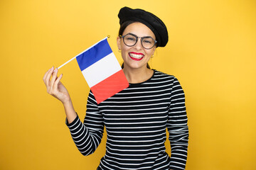 Young beautiful brunette woman wearing french beret and glasses over yellow background holding flag of French with a happy face standing and smiling with a confident smile showing teeth