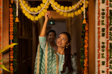 A YOUNG WOMAN HANGING SMALL LANTERN AT DECORATED ENTRANCE WITH HUSBAND LOOKING AT HER IN THE BACKGROUND	