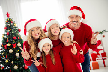 Photo portrait of funny friendly family wearing warm red festive jumpers and headwear preparing for christmas eve near xmas tree decorations showing v-sign with two fingers