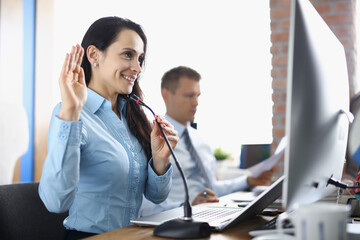 Smiling businesswoman sits at computer screen with microphone and waves hand with colleague in background in office. Remote business conference concept.