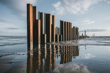 Large rusty steel construction pillars in the sand on a beach, with blue sky and calm waves. The steel girders are rusty. Building and construction on the coast. Development and building regulations.