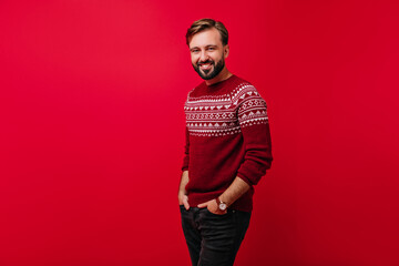 Refined man in wristwatch posing with hands in pockets on red background. Indoor shot of smiling brown-haired male model wears knitted norwegian sweater.