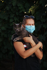 Woman in different poses in the garden with gray cat. She is wearing a safe mask to protect against infection.