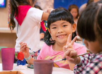 Portrait Asian child girl in art workshop, happy and smiling face, show gesture two fingers, cute face with black hair and bang. Blurred classmate foreground and background.
