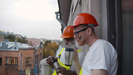 Diverse workers stand at balcony on construction site taking break and drinking coffee in thermos