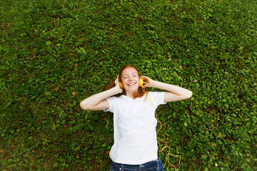 lifestyle, summer vacation, technology, music and the concept of people-a smiling young girl with red hair and headphones, lying with her eyes closed on the grass