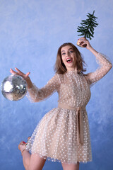 Naklejka premium beautiful happy woman in a beige dress holding a shiny disco ball and a small Christmas tree on a blue background. Holiday concept, new year, christmas, party.