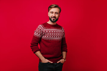 Confident white man with beard posing with hands in pockets. Studio shot of handsome guy with dark hair wears norwegian sweater. - obrazy, fototapety, plakaty